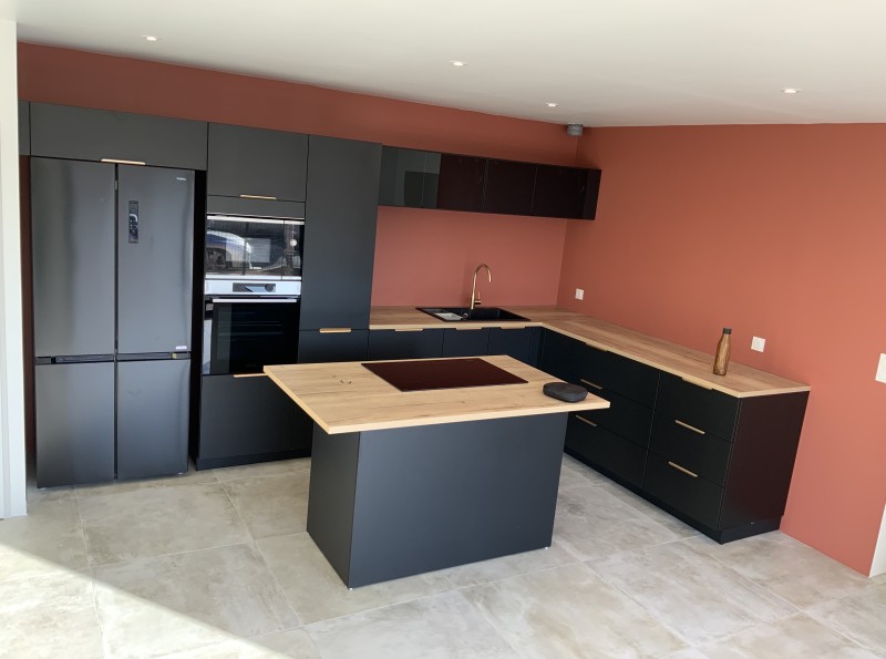 Création d'une cuisine en L noire mate avec îlot cuisson et plans bois sur mur terracotta à Lunel